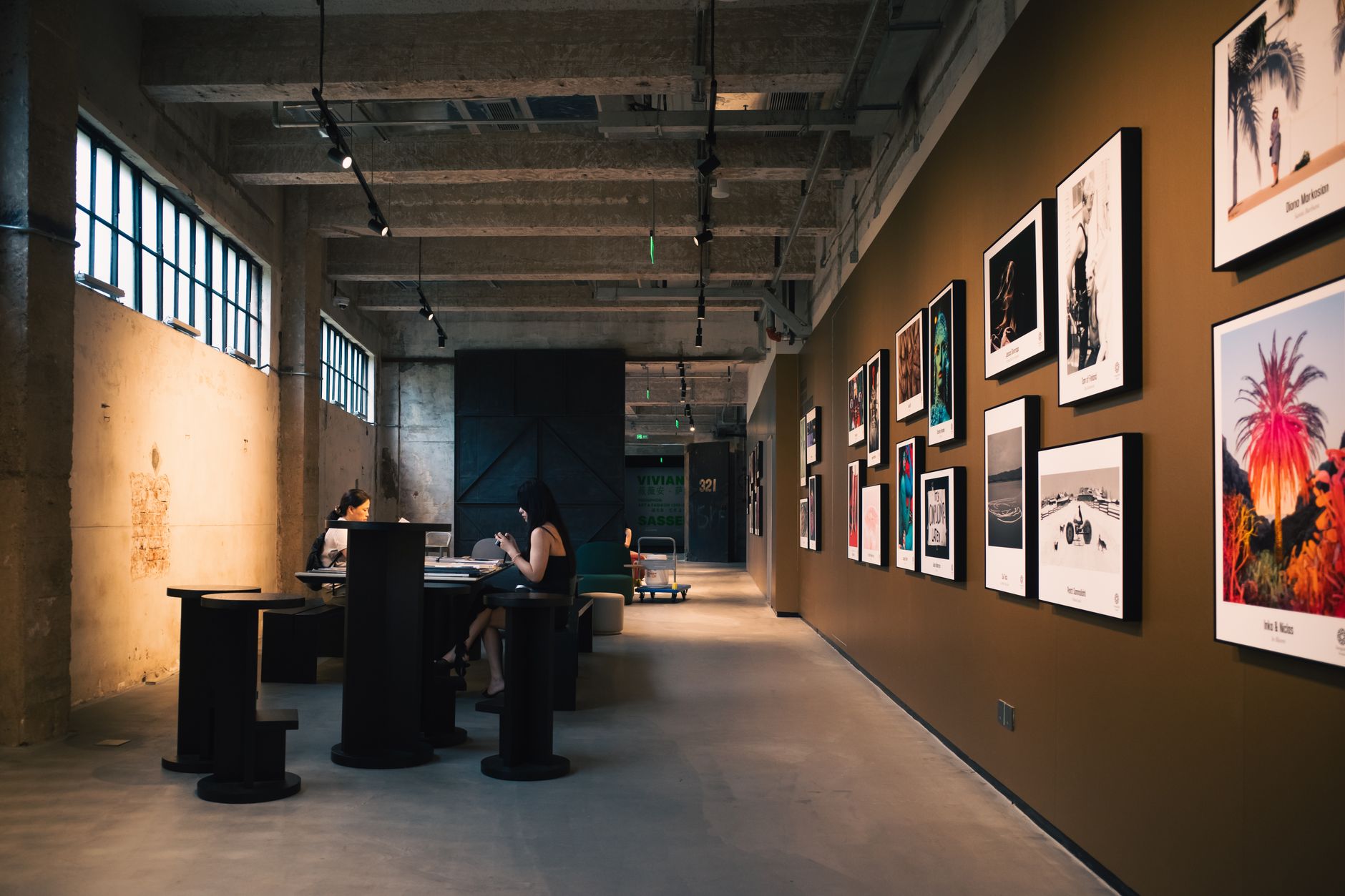 Art gallery hallway with framed artworks and visitors seated inside, under warm lighting.