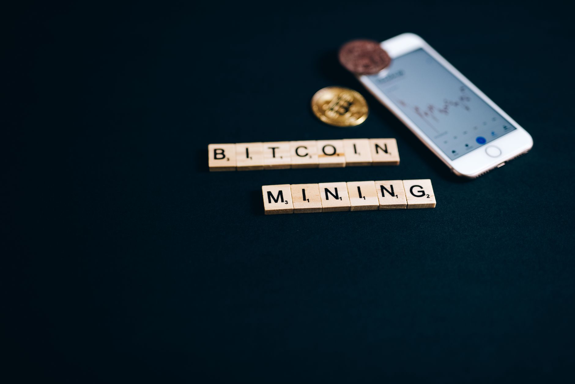 Scrabble tiles spelling Bitcoin Mining with smartphone and coins on a dark background.