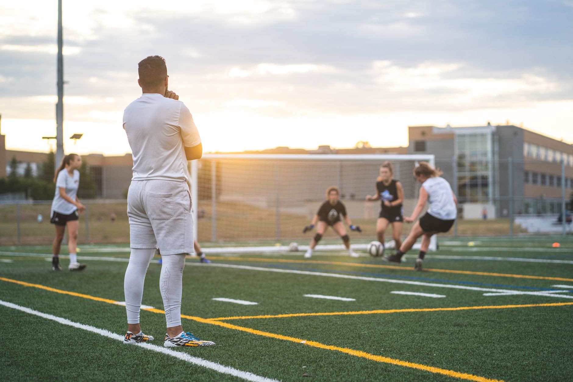 Group of women with coach training soccer together on sports ground with lines under cloudy sunny sky in summer in back lit