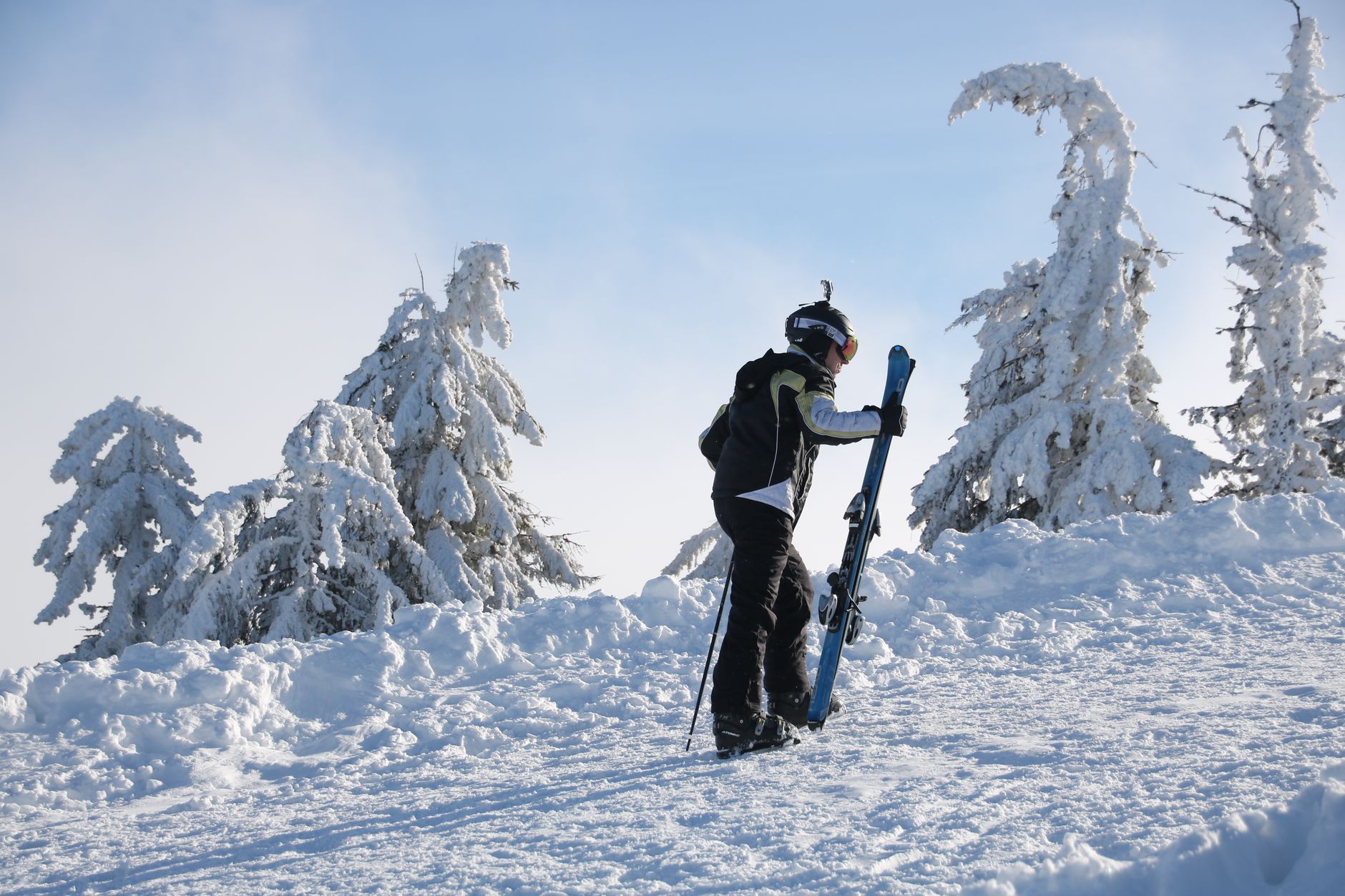 A lone skier carries equipment uphill through a snow-laden landscape, dressed in winter sports gear.