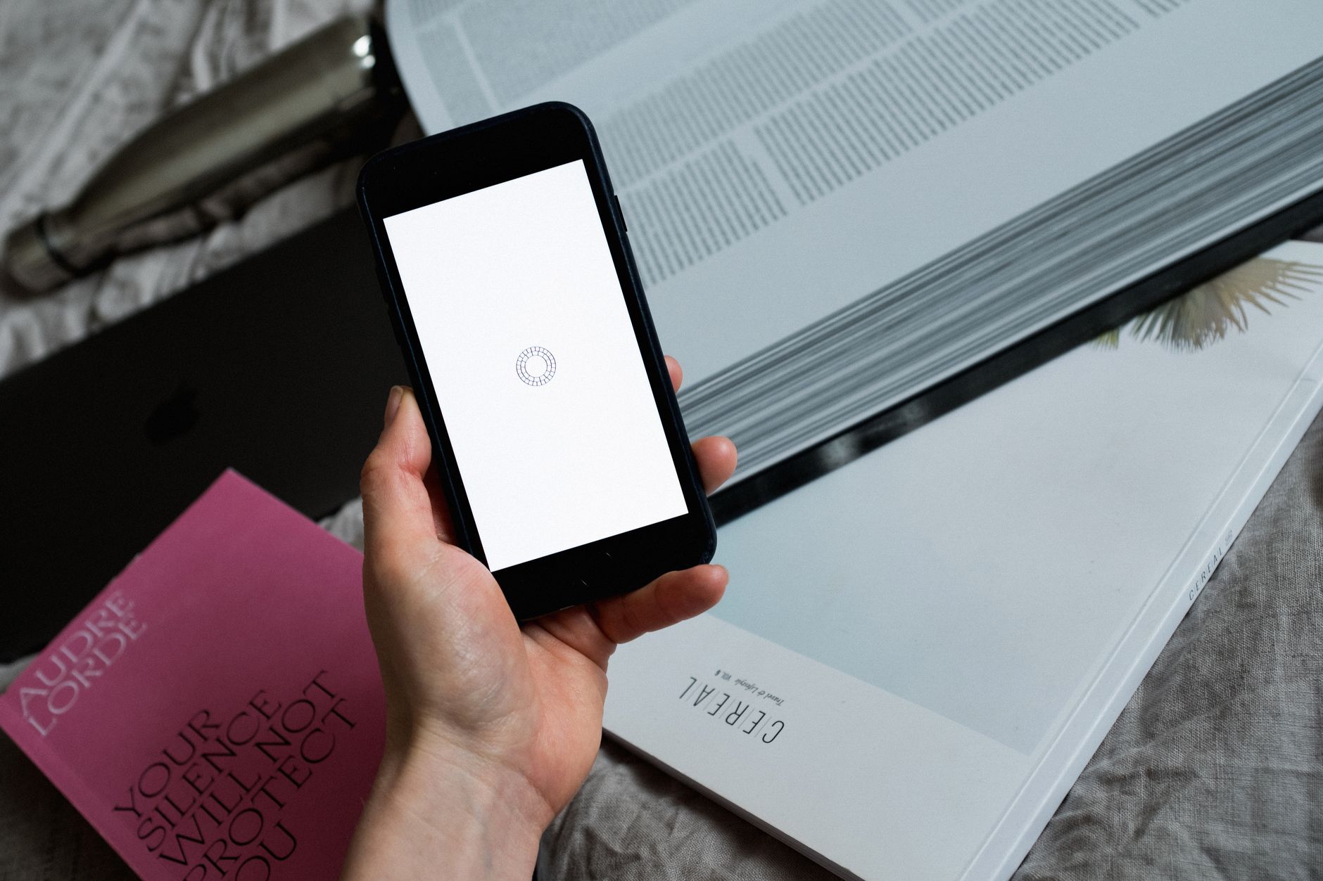 From above of crop anonymous person demonstrating mobile phone blank screen with loading symbol while having gadget near open book located on sheet
