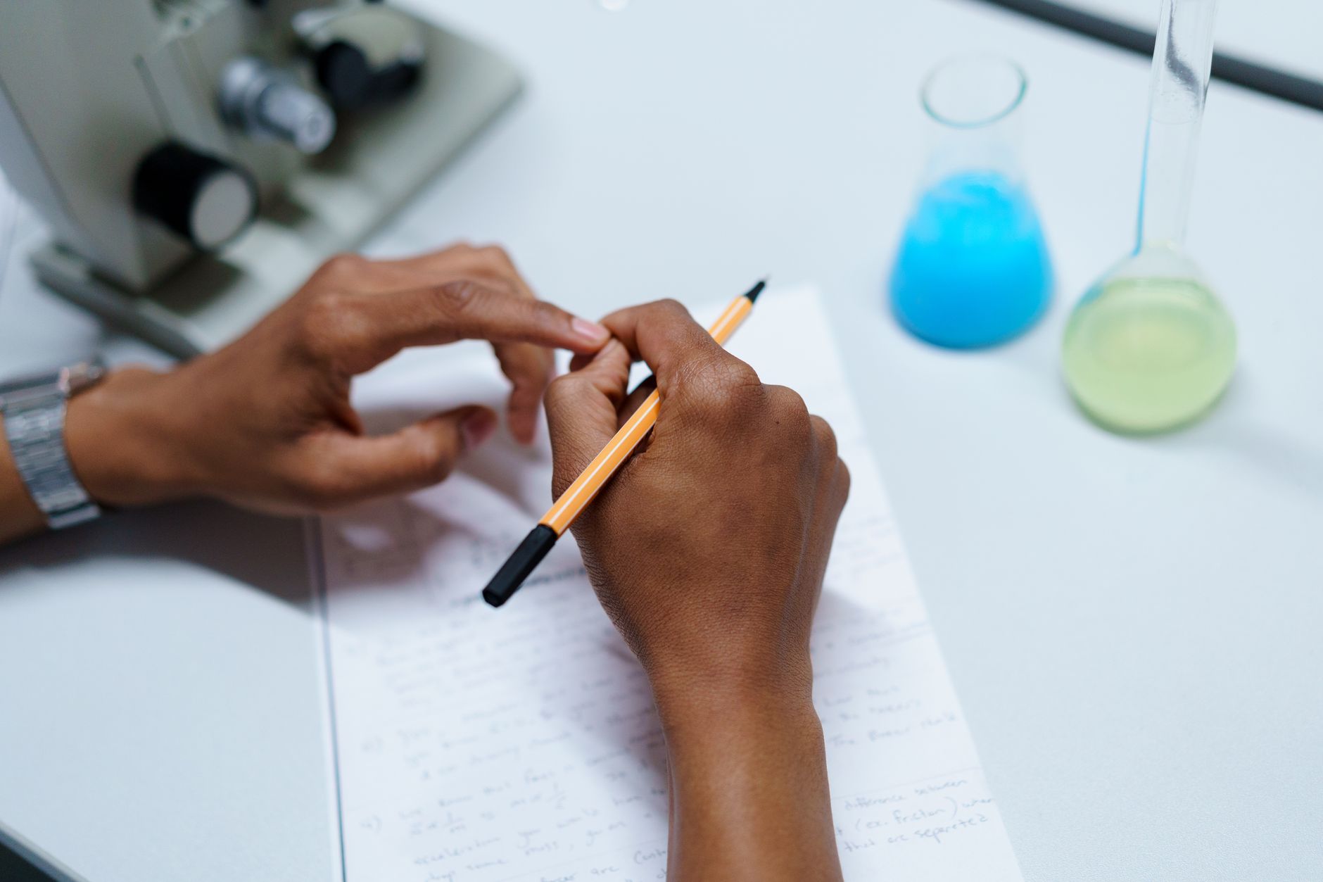 Close-up of a scientist taking notes in a laboratory setting with flasks and a microscope.