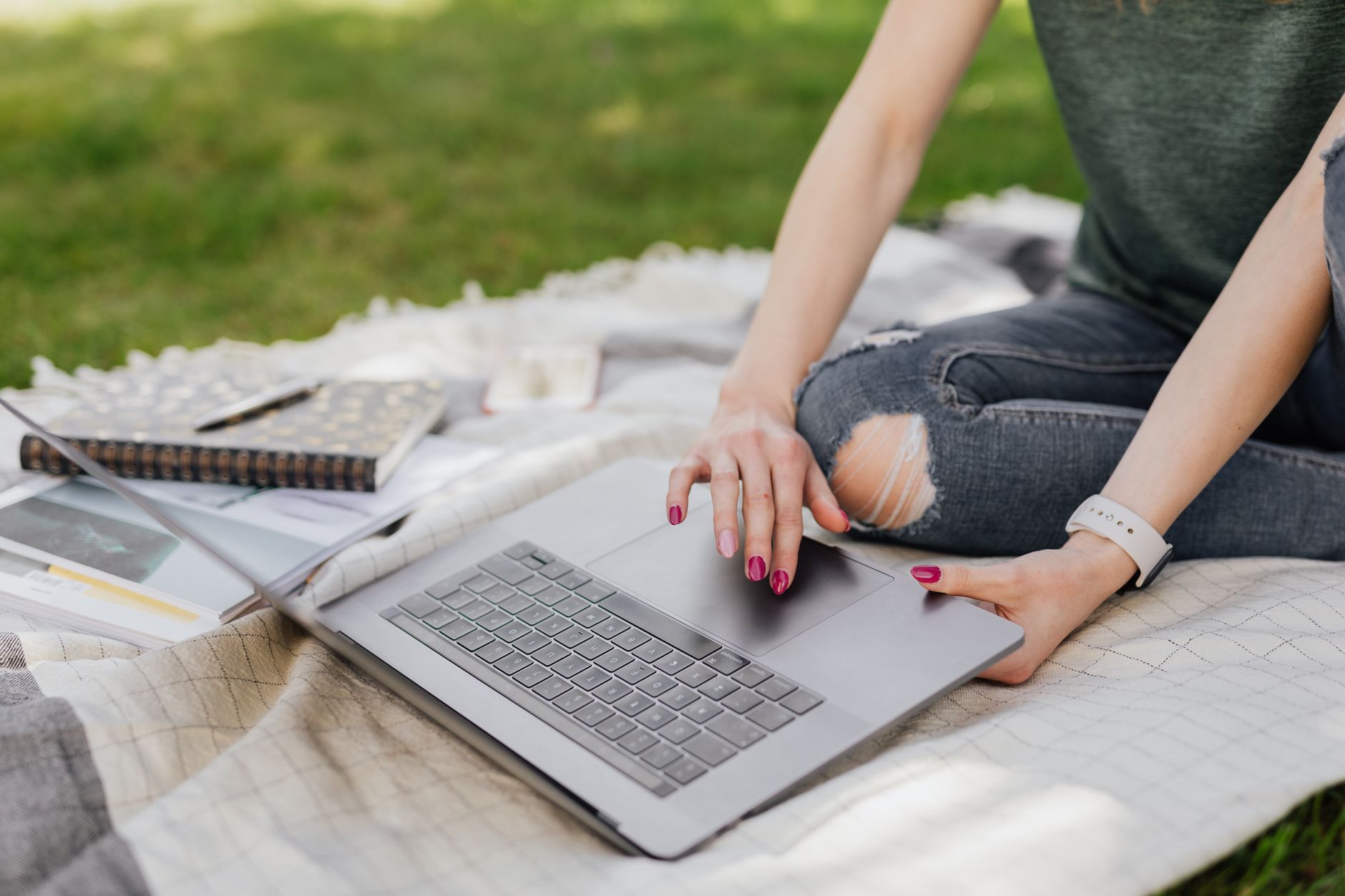 Crop unrecognizable female student doing Internet research on laptop while sitting on cozy blanket in green summer park on sunny day