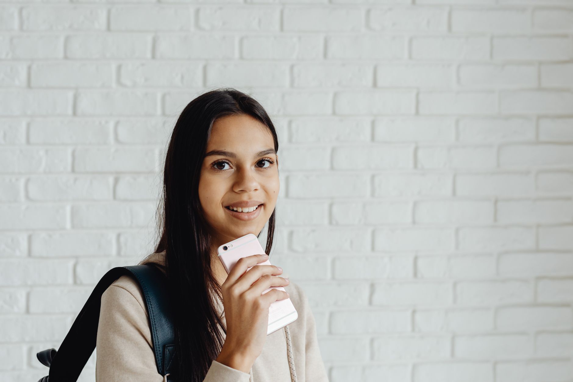 A young woman with black hair holds a smartphone while smiling indoors.