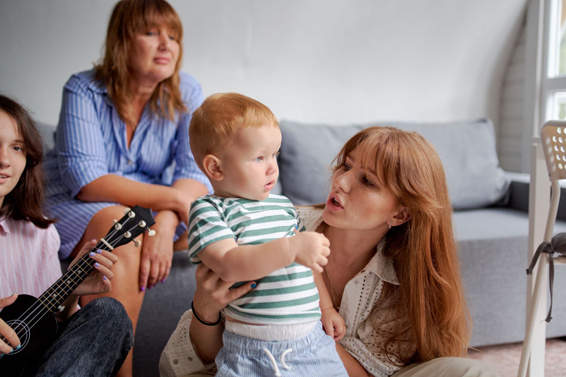 Women with toddler child near crop grandmother and sister playing ukulele while spending time together on weekend