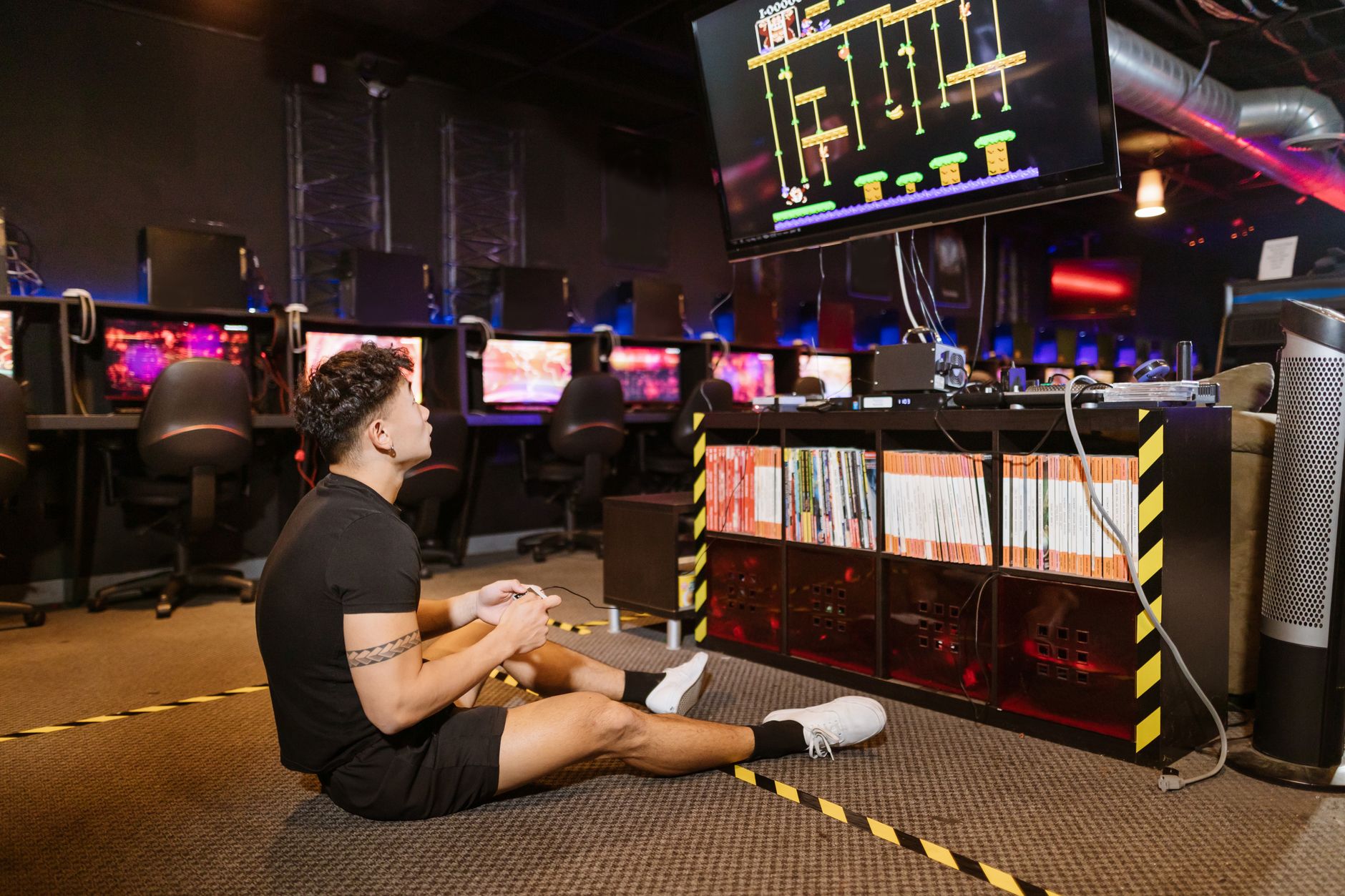 Man sitting on floor intensely playing video game at a modern arcade with vibrant computing equipment.