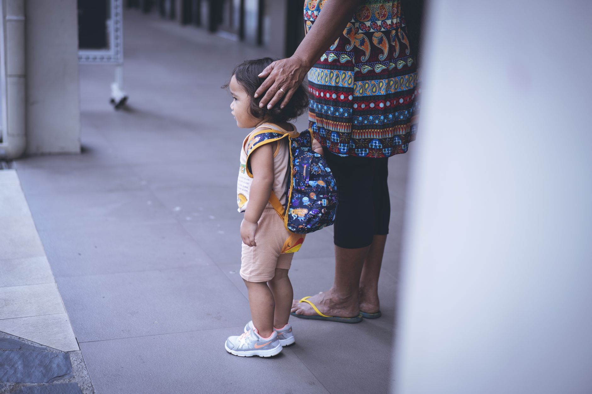 A young child with a backpack stands beside a caregiver's protective touch outdoors.