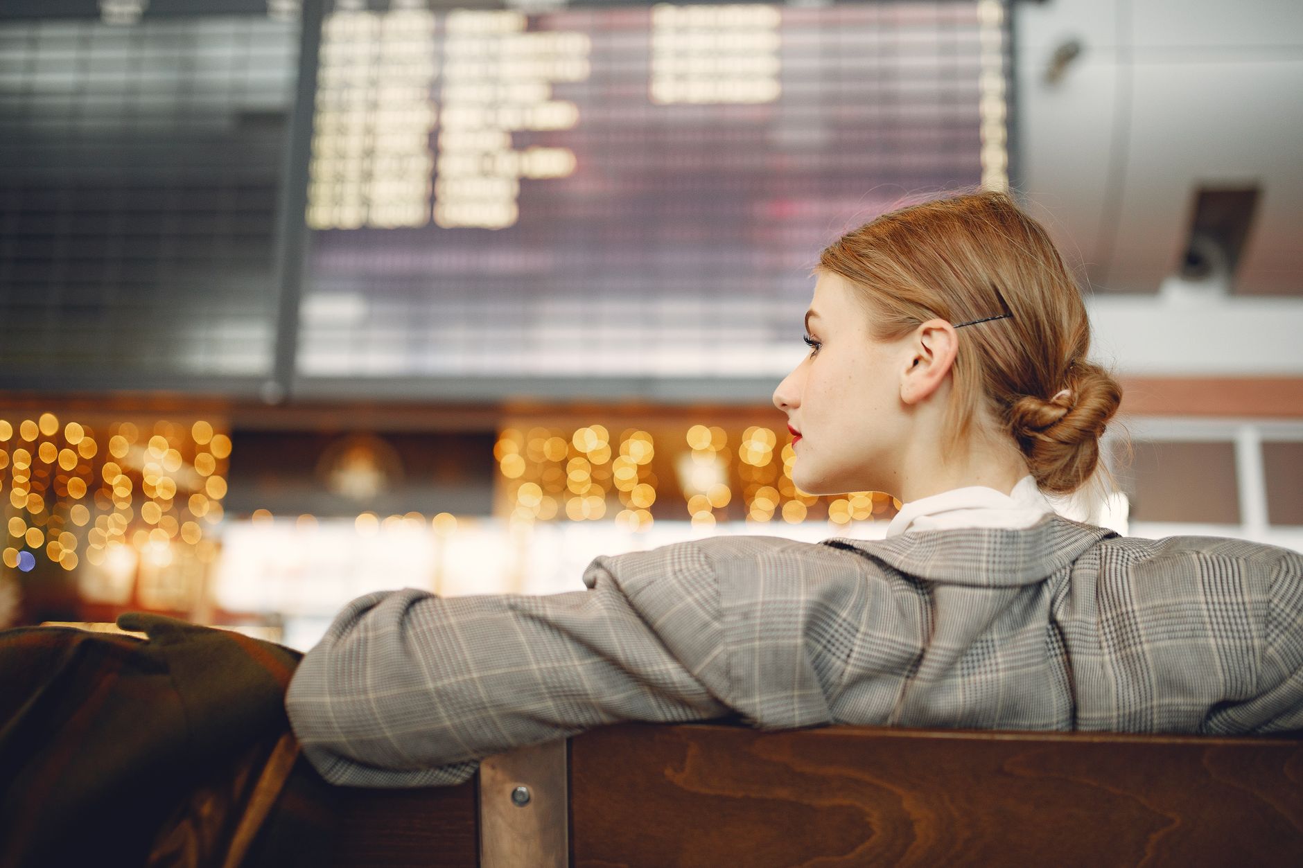 Pensive female designer in checkered jacket waiting for flight near departure board looking away in modern airport in evening