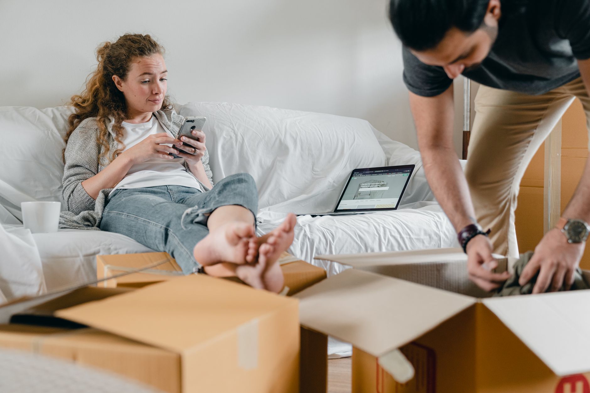 Barefoot female in casual wear sitting with crossed legs on sofa with open laptop while using social media on cellphone and crop male partner getting clothes out of cardboard box
