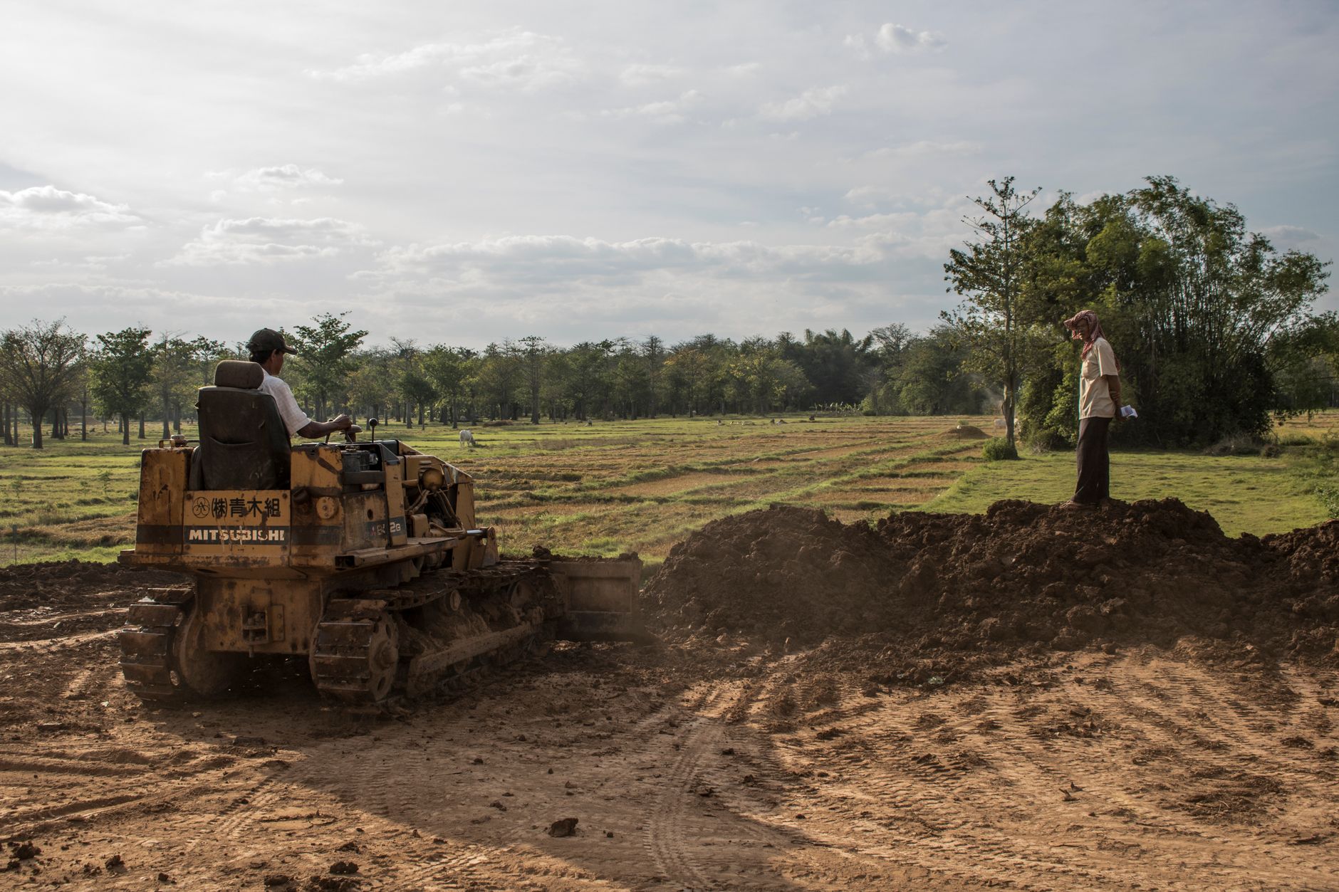 Excavator working on a rural field with workers, surrounded by greenery.