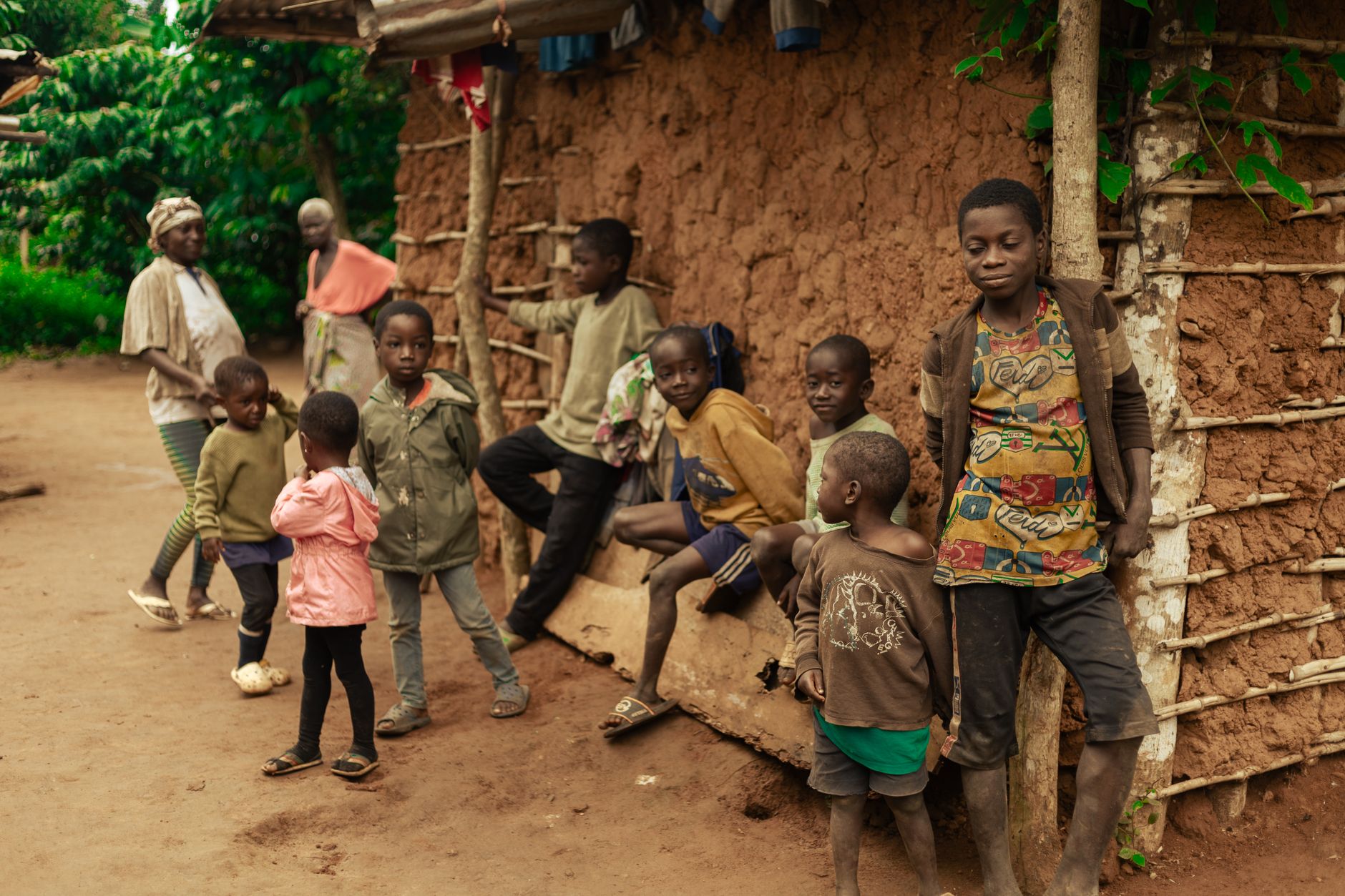 A group of children and adults gather outside a traditional mud house in a vibrant village setting.