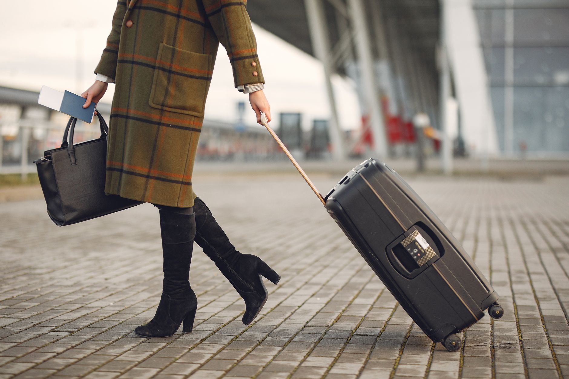 Crop young female traveler in trendy plaid coat and high boots with luggage and documents strolling on pavement near contemporary building of airport during cold season