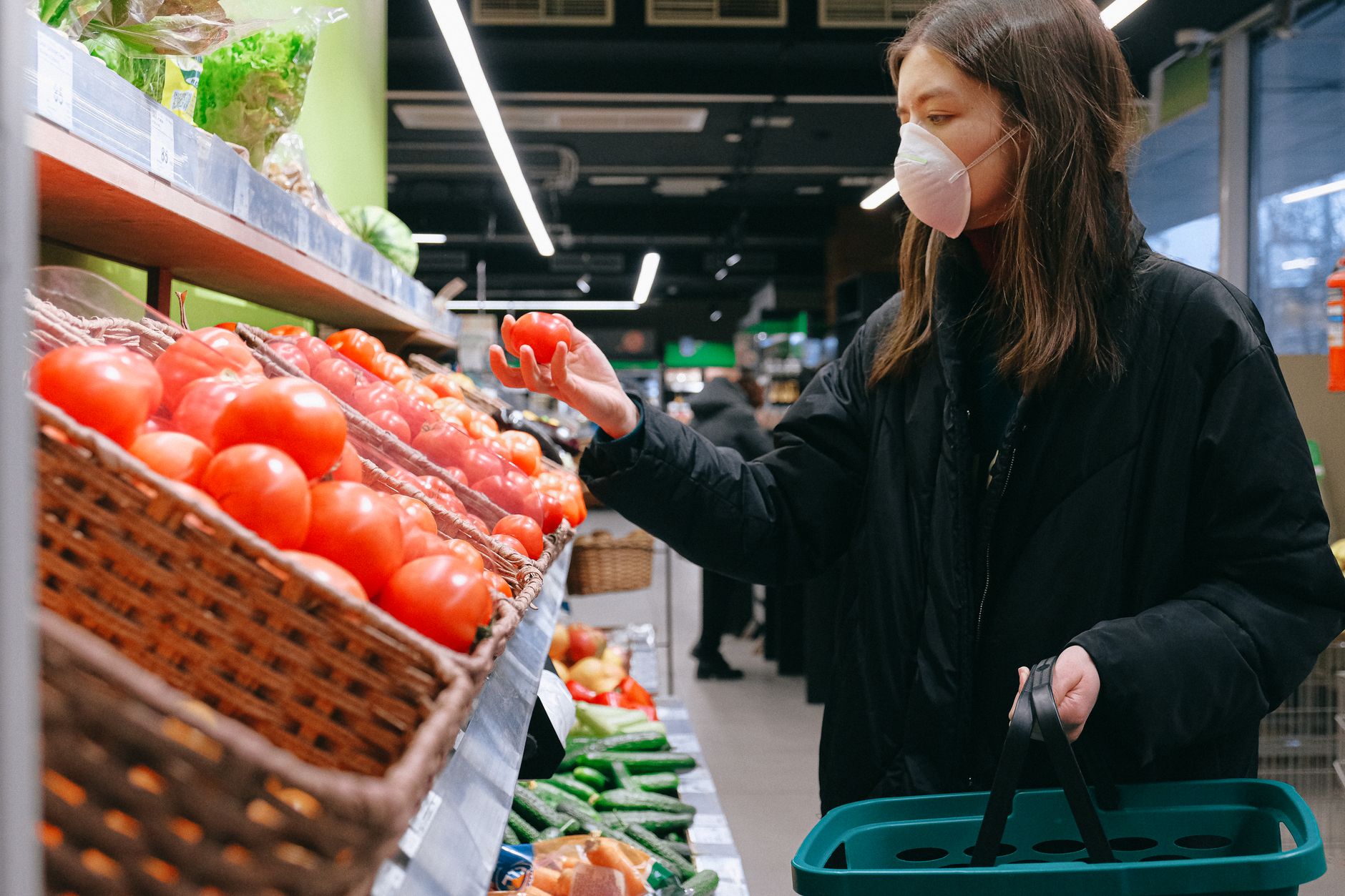 A woman in a face mask selects tomatoes at a grocery store, showcasing pandemic shopping habits.