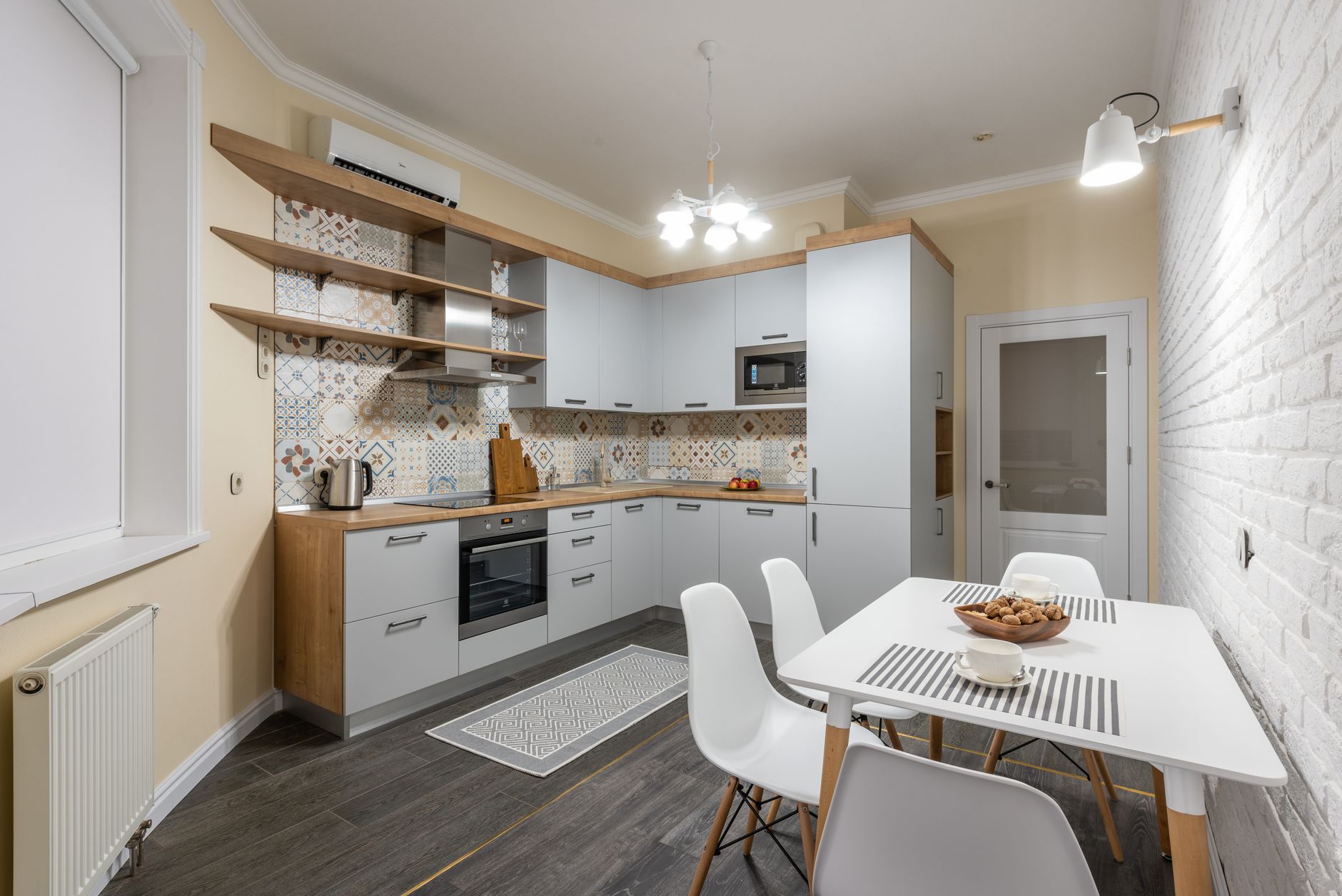 Modern kitchen with electric appliances and radiator against table with tasty biscuits and cups illuminated by bright chandelier