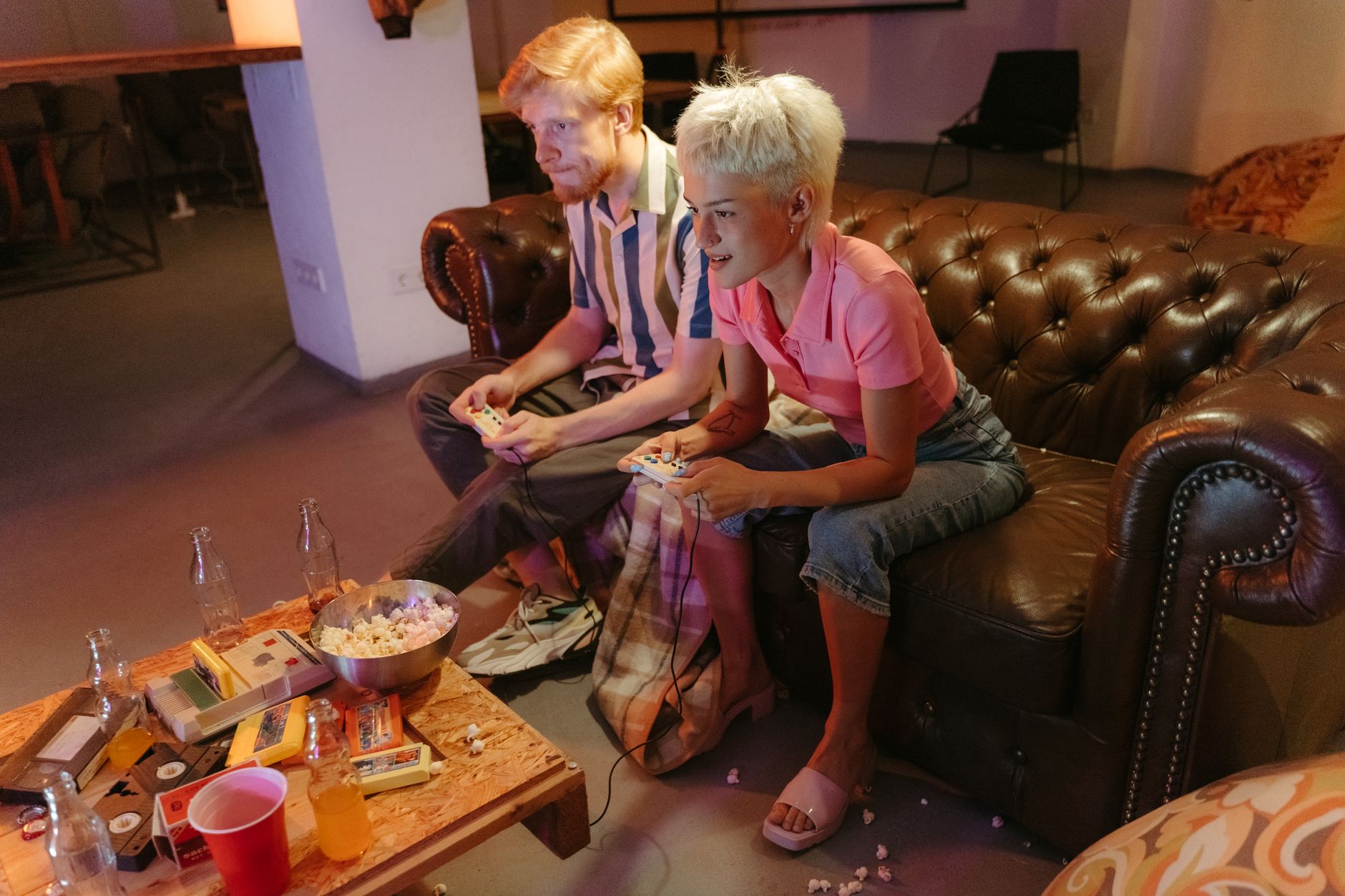 Two friends playing video games on a leather sofa, surrounded by snacks and drinks, enjoying a casual gaming night.