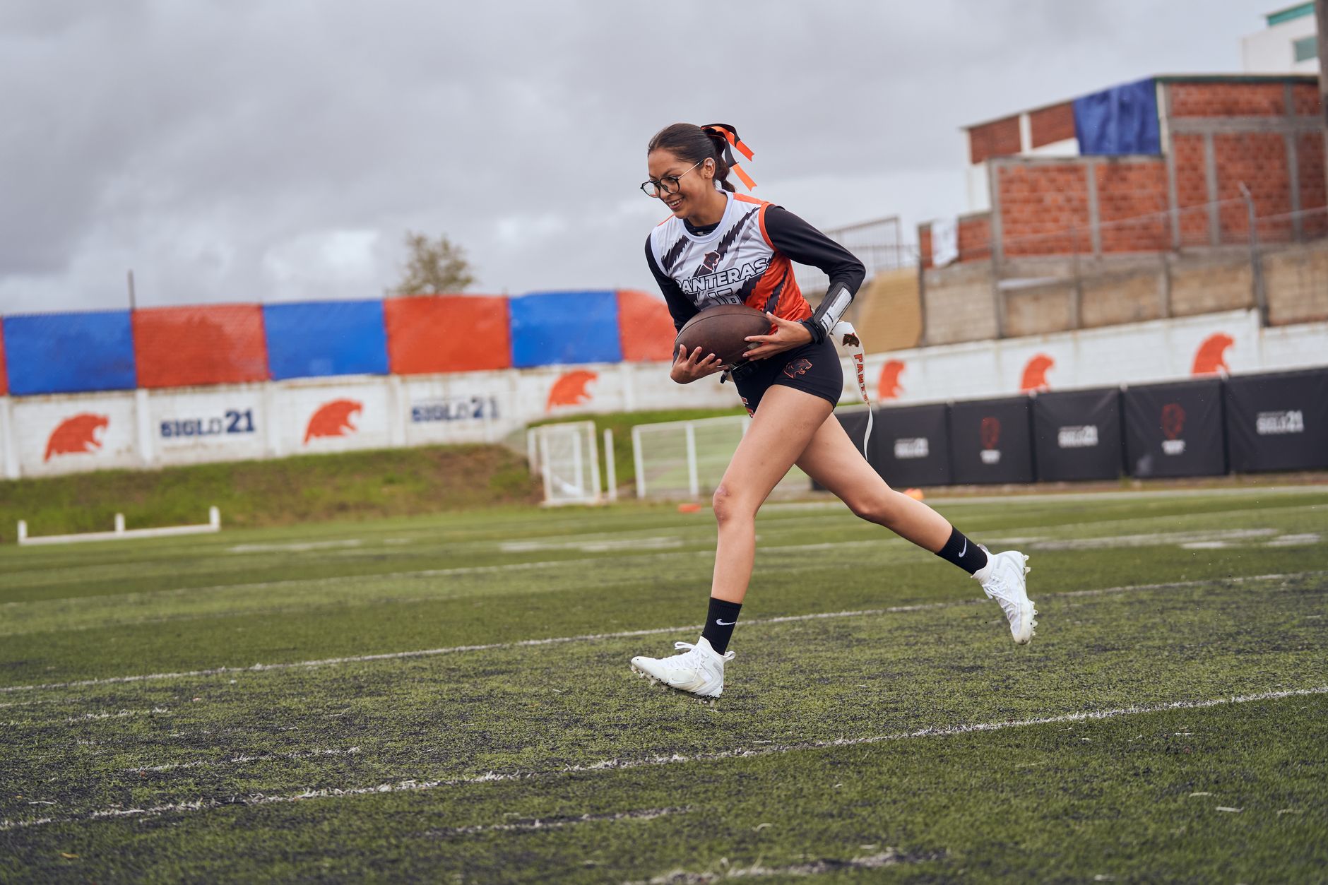 Female athlete playing American football on a grass field during practice under cloudy skies.