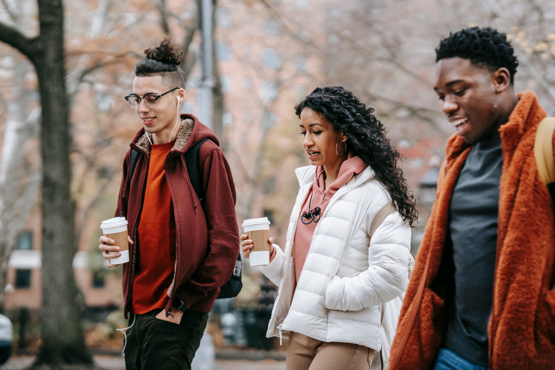 Confident young multiracial friends in outerwear and backpacks talking while strolling in city street near trees with paper cups with drink and earphones in daytime in park