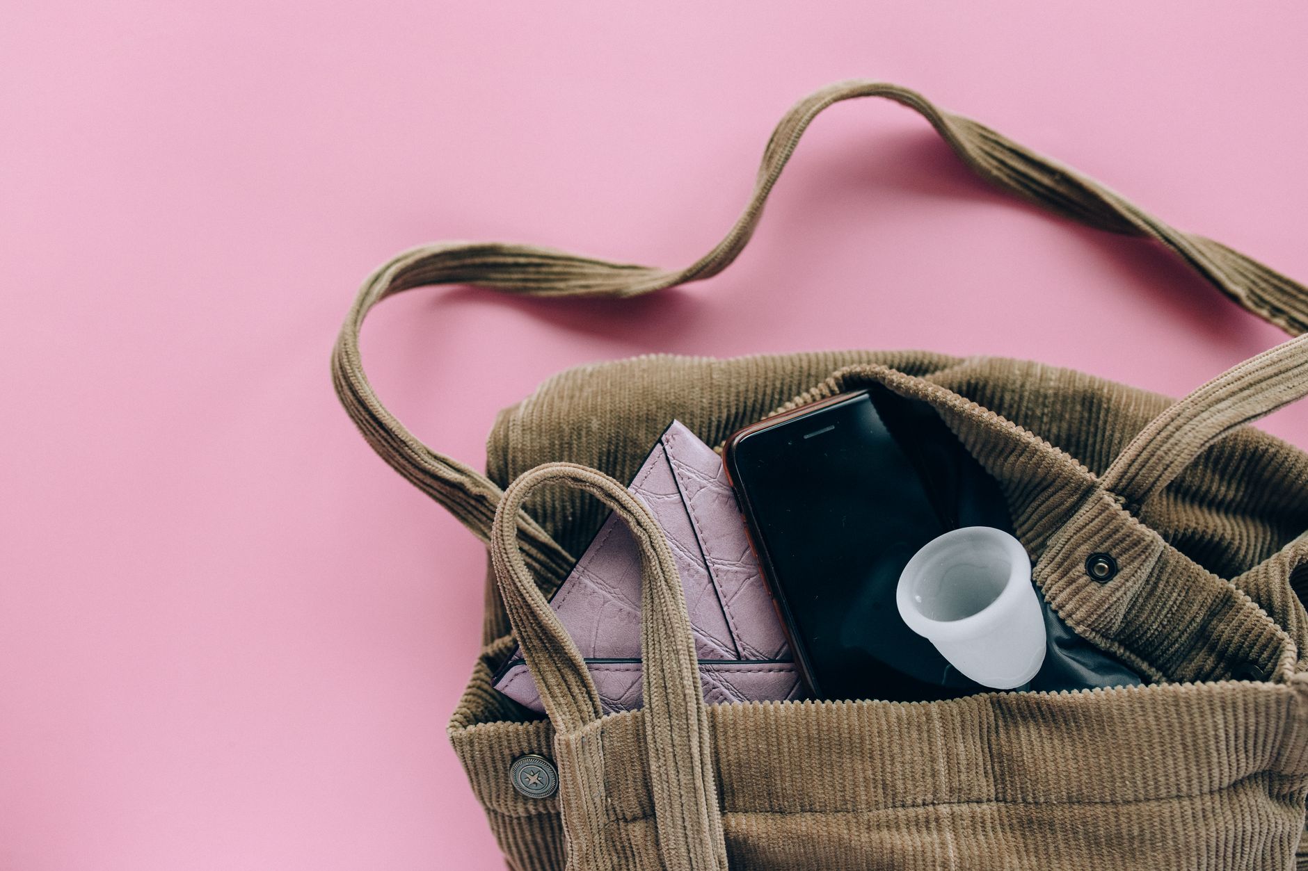 Close-up of a corduroy bag containing a smartphone and menstrual cup against a pink background.