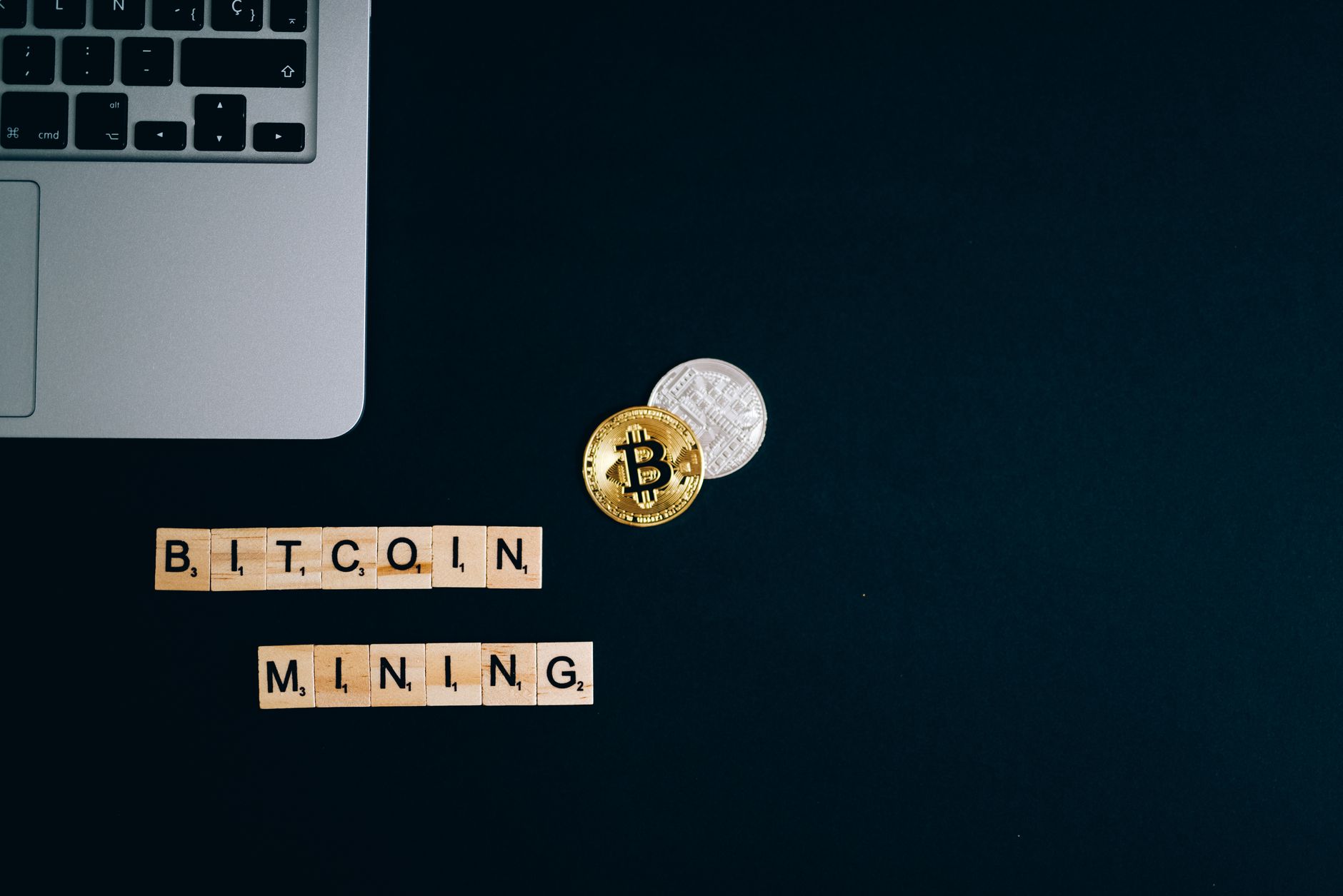 Top view of a laptop with Bitcoin coins and blocks spelling 'Bitcoin Mining' on a dark surface.