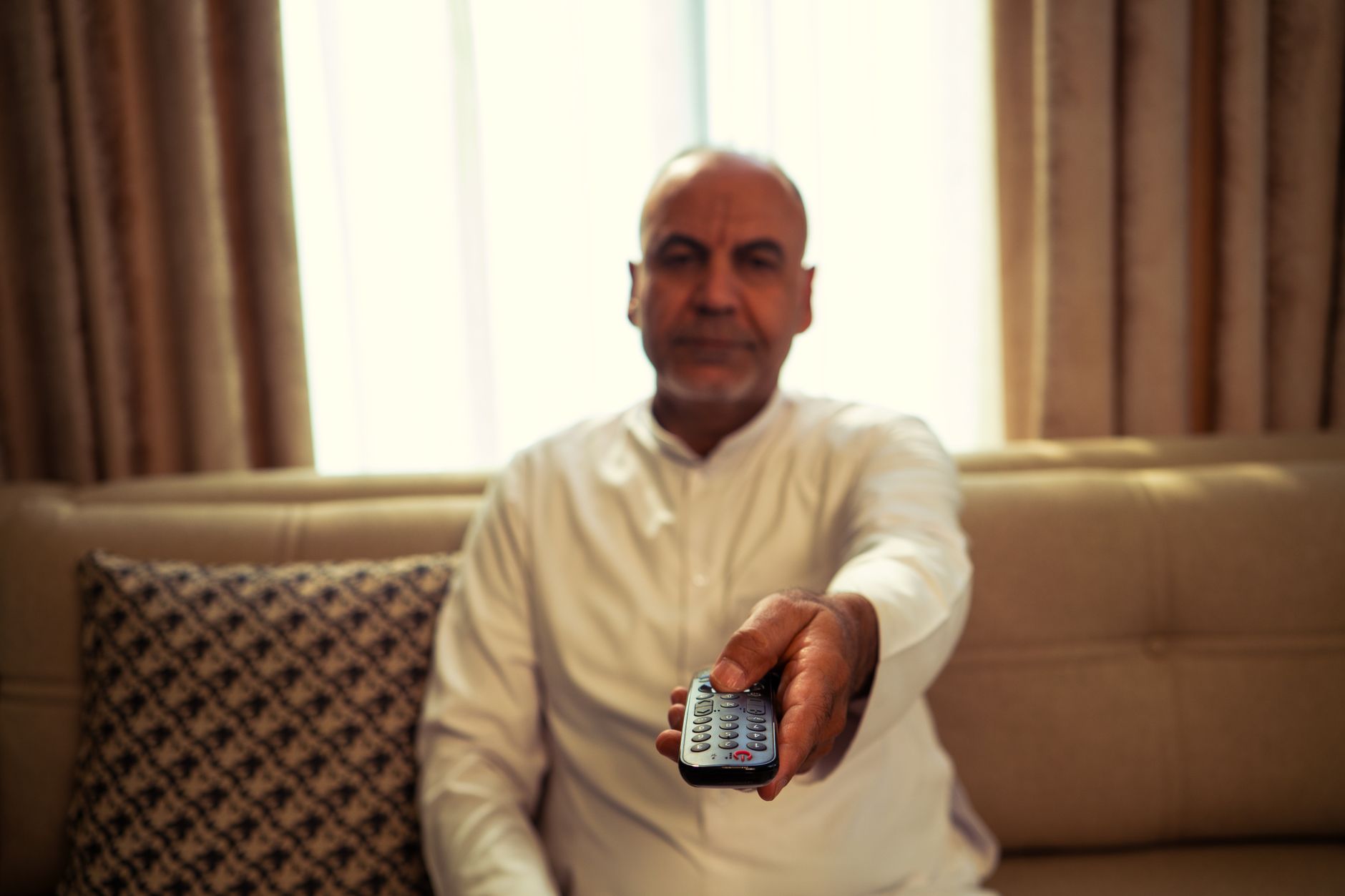 A senior man in a living room extends a TV remote towards the camera.