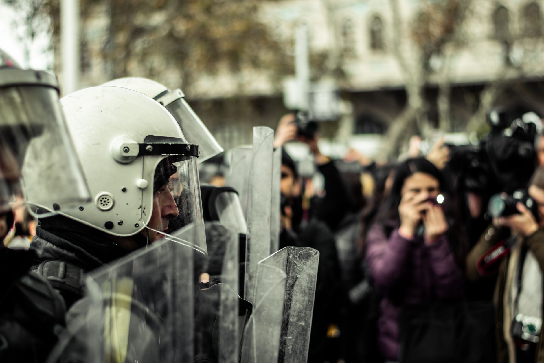 Police officers in helmets and shields confronted by crowd of photographers.