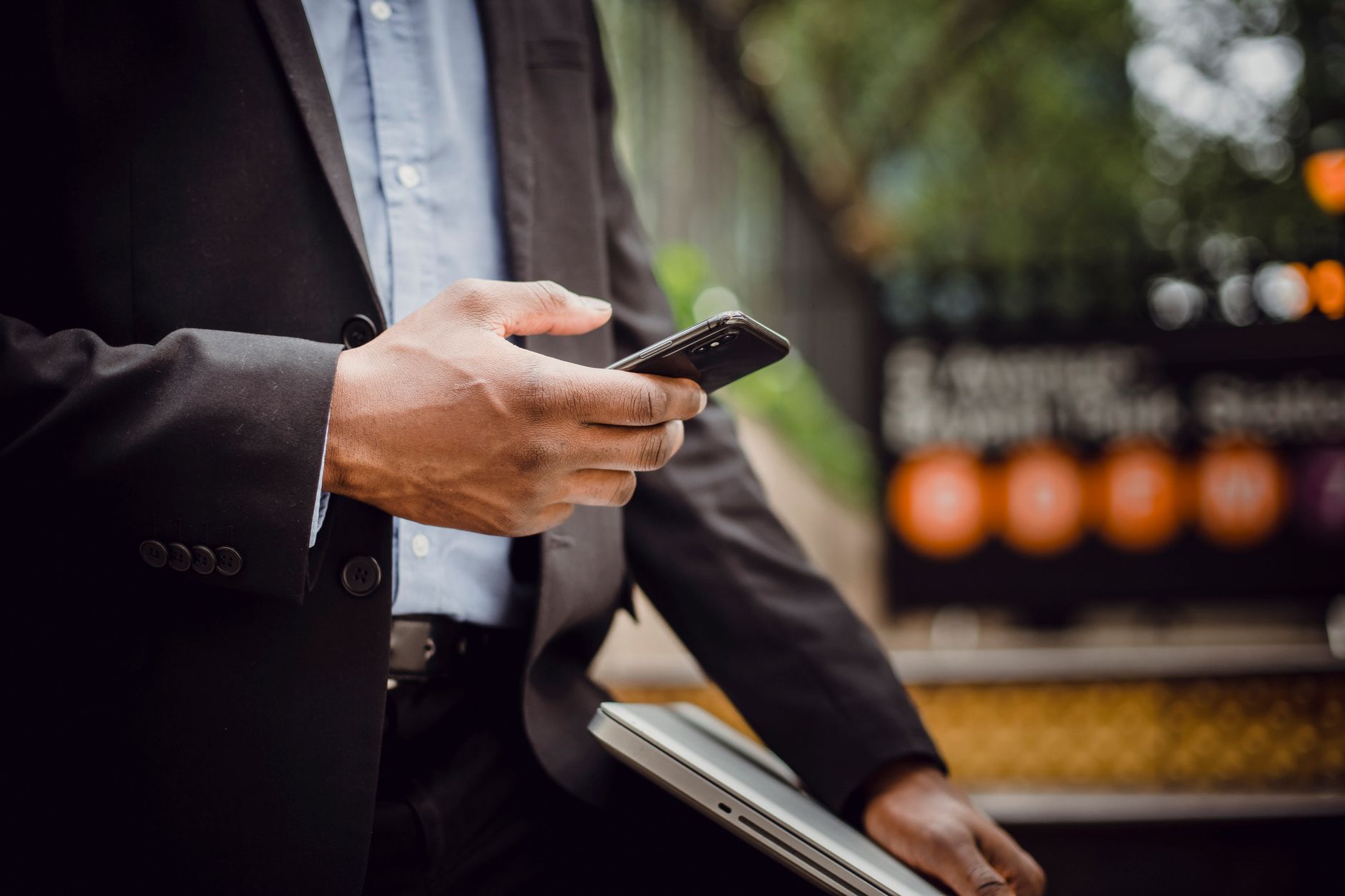 Crop unrecognizable African American man in formal wear browsing mobile phone while standing with laptop on street during sunny day